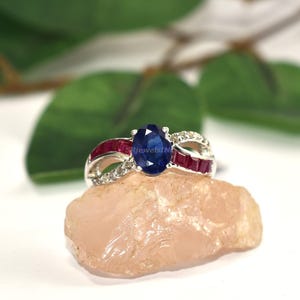 Peut inclure: Une bague en argent ornée d'une pierre précieuse ovale bleu foncé au centre, flanquée de pierres rouges rectangulaires et de petits diamants blancs. La bague est posée sur une pierre rose clair, avec des feuilles vertes en arrière-plan.