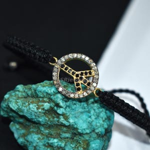 May include: A black cord bracelet featuring a gold-toned peace symbol pendant. The pendant is encrusted with black and clear gemstones. The bracelet is displayed on a turquoise stone against a black and white background.