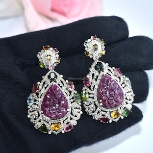 Peut inclure: Une paire de boucles d'oreilles pendantes ornées. Chaque boucle d'oreille présente une grande pierre précieuse rose foncé sculptée au centre, entourée de pierres précieuses colorées plus petites et de cristaux transparents. Les boucles d'oreilles ont un design ajouré détaillé avec un sertissage en métal argenté.