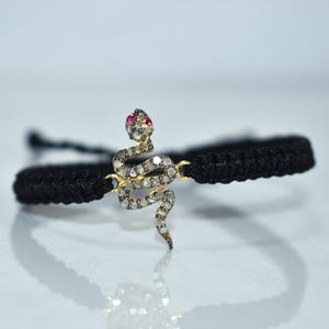 May include: A black woven bracelet with a gold-coloured snake charm. The snake is adorned with small, clear stones and a single red stone at the head. The bracelet is shown on a reflective surface.