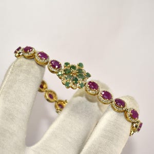 May include: Gold bracelet with oval-cut ruby gemstones and emeralds. The bracelet has a floral design with a cluster of emeralds and diamonds. The rubies are set in gold with diamond accents, creating a luxurious appearance.