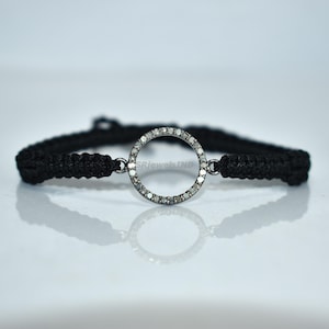 May include: A black macrame bracelet featuring a circular silver pendant encrusted with small, sparkling stones. The bracelet's cord is tightly woven, and the pendant is centered, creating a minimalist and elegant design. The bracelet is suitable for everyday wear.