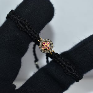 Könnte beinhalten: Schwarz geflochtenes Armband mit einem goldfarbenen, kreisförmigen Mittelstück. Das Mittelstück hat ein Blumenmuster mit rosa Edelsteinen und ist von kleinen, klaren Steinen umgeben. Das Armband wird von einer behandschuhten Hand gehalten.