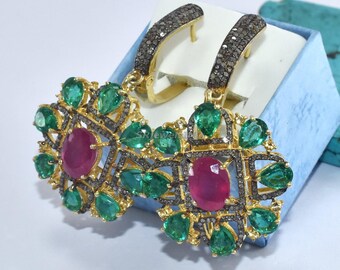 May include: A pair of ornate earrings featuring a central oval ruby surrounded by emeralds and small black stones. The earrings have gold-colored hoops and are displayed in a blue box.