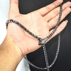 Puede incluir: Un collar gris oscuro con un diseño geométrico. El collar presenta un colgante central con un patrón de diamantes. El collar se sostiene en una mano sobre un fondo oscuro.