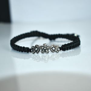 May include: Black braided bracelet with a floral design. The bracelet features a series of three flower-shaped charms, each adorned with small, sparkling stones. The bracelet is set against a white background.