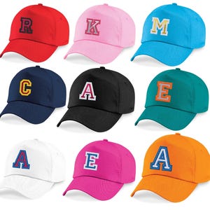 May include: A collection of nine baseball caps in various solid colours including red, pink, light blue, navy, black, teal, white, magenta, and orange. Each cap features a large, embroidered collegiate-style letter on the front panel.