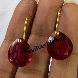May include: A pair of gold-colored drop earrings with large, faceted, teardrop-shaped, red gemstones. The earrings are displayed against a neutral background. The text "Thesilverhiphop" is visible on the image.