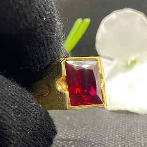 Könnte beinhalten: Ein goldener Ring mit einem rechteckigen, tiefroten Edelstein. Die Fassung des Rings ist hellgold, und der Edelstein ist der Mittelpunkt. Der Ring wird von einer schwarz behandschuhten Hand gehalten, mit einer weißen Blume im Hintergrund.