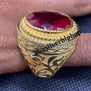 Könnte beinhalten: Ein goldfarbener Ring mit einem großen, ovalen, roten Edelstein. Der Ring hat aufwendige eingravierte Muster und kleine, klare Steine um den Edelstein herum. Der Text "thesilverhiphop" ist auf dem Ring sichtbar.