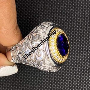 Può includere: Anello in argento con una gemma ovale blu scuro, circondata da una fascia color oro e piccole pietre bianche. L'anello presenta un design ornato e inciso sulla fascia. Il testo "thesilverhiphop" è visibile.