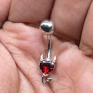 Puede incluir: Un piercing para el ombligo plateado con una gema roja en forma de corazón, cuernos de diablo y una cola. La bola superior también es plateada. Esta joya corporal única añade un toque de estilo atrevido.