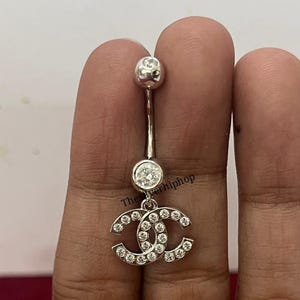 May include: A silver belly button ring featuring a double-C logo design encrusted with clear crystals. The top and bottom balls are also adorned with crystals. The jewelry is held up by a slender silver bar.