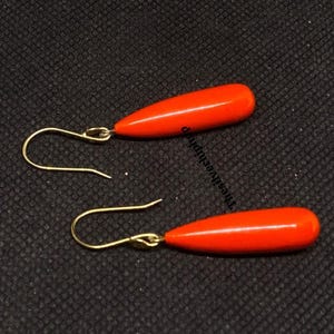 Può includere: Un paio di orecchini arancioni a goccia con ganci dorati. Gli orecchini hanno una finitura liscia e lucida e sono su uno sfondo scuro.