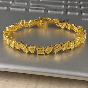Op de afbeelding: Een goudkleurige armband met een reeks gele, verschillend gevormde edelstenen, waaronder peer-, marquise-, vierkante en smaragdgeslepen stenen. Het sieraad heeft een bakslot en de tekst Thesilverhiphop is zichtbaar bij de sluiting.