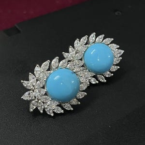 May include: A pair of silver stud earrings. Each earring has a large, round, light blue stone, encircled by smaller, clear, marquise-cut stones. The earrings are displayed on a dark surface.