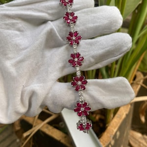 Puede incluir: Una pulsera de tono plateado que presenta una serie de seis motivos en forma de flor. Cada flor se compone de seis piedras de color rosa intenso en forma de lágrima dispuestas alrededor de una piedra central clara, con pequeñas piedras claras uniendo cada eslabón.