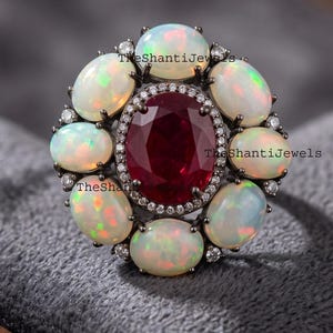 May include: A dark metal ring featuring a large oval-cut deep red center stone surrounded by a halo of small white accent stones. Eight oval cabochon opals with iridescent flashes encircle the center, set in a dark metal band with text.