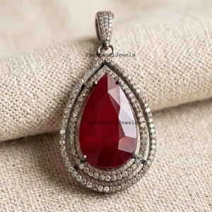 May include: A silver-toned pendant with a pear-shaped, faceted, deep red gemstone at its centre. The gemstone is framed by two rows of small, sparkling stones. The pendant has a textured bail. The text "TheShantiJewels" is visible on the pendant.