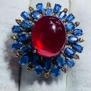 May include: A ring featuring a large, oval, cabochon-cut deep red gemstone at the center, surrounded by a halo of smaller, oval-cut blue sapphires and tiny, round, clear accent stones set in a metallic frame with a polished finish.
