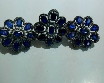May include: A jewellery set consisting of three flower-shaped pieces crafted from dark metal. Each piece features a central oval blue gemstone surrounded by a ring of smaller oval blue gemstones, all accented with tiny clear pavé stones.