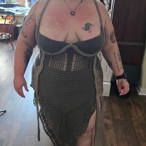Może przedstawiać: Ręcznie robiona, szara sukienka z szydełka z topem typu halter i sp&oacute;dnicą z frędzlami. Sukienka ma unikalny design z dopasowanym gorsetem i asymetrycznym dołem. Osoba ma na sobie srebrne chodaki.