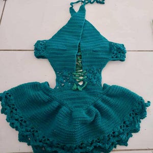 May include: Teal crochet halter dress with a flared skirt and decorative edging. The dress features a laced bodice and short sleeves. The halter straps are tied at the neck. The dress is handmade and has a textured appearance.