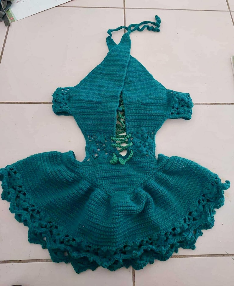 May include: Teal crochet halter dress with a flared skirt and decorative edging. The dress features a laced bodice and short sleeves. The halter straps are tied at the neck. The dress is handmade and has a textured appearance.