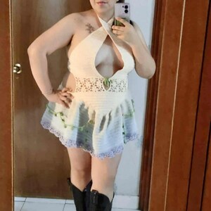 May include: A cream-coloured, handmade crochet dress with a halter neck and flared skirt. The dress features a decorative lace detail at the waist and a blue and green trim. The outfit is completed with black cowboy boots.
