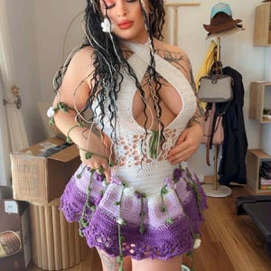 May include: A handmade crochet outfit featuring a white halter top with a keyhole design and a purple and white skirt. The outfit is adorned with green vines and white flowers. The model has long braids with floral accents.