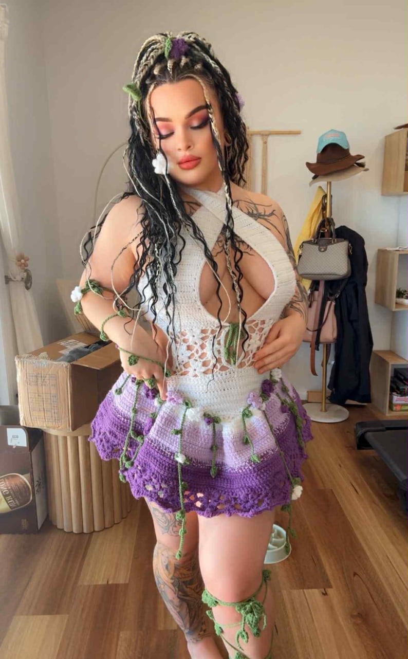 May include: A handmade crochet outfit featuring a white halter top with a keyhole design and a purple and white skirt. The outfit is adorned with green vines and white flowers. The model has long braids with floral accents.