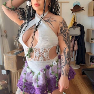 May include: A white halter top and a purple crochet skirt with floral and vine accents. The outfit is handmade and features a unique design. The model is wearing accessories that match the outfit.