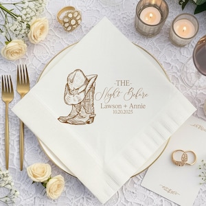 May include: A white paper napkin featuring a brown line-art illustration of a cowboy hat and boots. The text reads THE Night Before, Lawson + Annie, and 10.20.2025. The napkin is square with a textured border and is placed on a white plate.