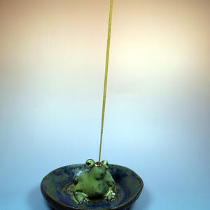 May include: A ceramic incense holder featuring a green frog figurine in a blue dish. The frog has detailed features and sits in the center of the dish. A lit incense stick stands upright, emitting a thin stream of smoke.
