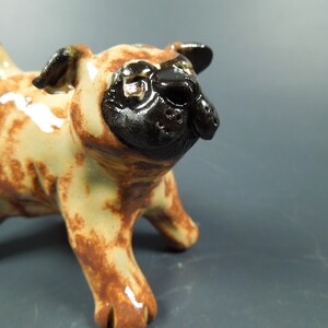 Puede incluir: Una peque&ntilde;a figura de cer&aacute;mica de un perro pug con acabado brillante. El cuerpo es de color tostado claro con manchas marrones irregulares, mientras que la cara, las orejas y la nariz son de un color negro s&oacute;lido. El pug est&aacute; de pie mirando al frente.