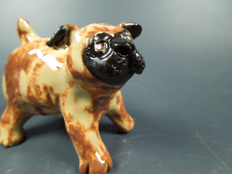 Puede incluir: Una peque&ntilde;a figura de cer&aacute;mica de un perro pug con acabado brillante. El cuerpo es de color tostado claro con manchas marrones irregulares, mientras que la cara, las orejas y la nariz son de un color negro s&oacute;lido. El pug est&aacute; de pie mirando al frente.