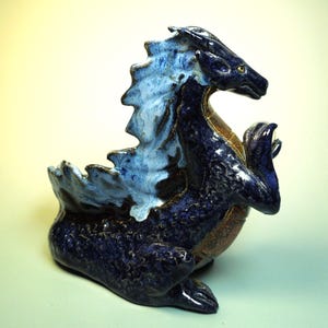 May include: A ceramic dragon figurine in shades of blue and brown. The dragon has a textured, scaled body, with a light blue and white frill along its back. The figurine is posed in a seated position with its claws raised.
