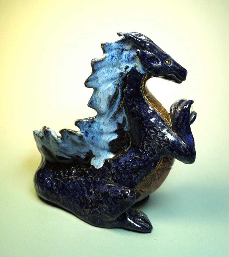 May include: A ceramic dragon figurine in shades of blue and brown. The dragon has a textured, scaled body, with a light blue and white frill along its back. The figurine is posed in a seated position with its claws raised.