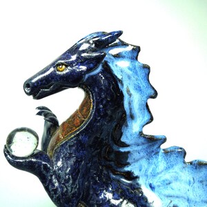 May include: A ceramic dragon figurine in shades of blue, holding a clear glass orb. The dragon has a detailed, textured surface with a contrasting orange-brown chest. The figurine is a decorative item.