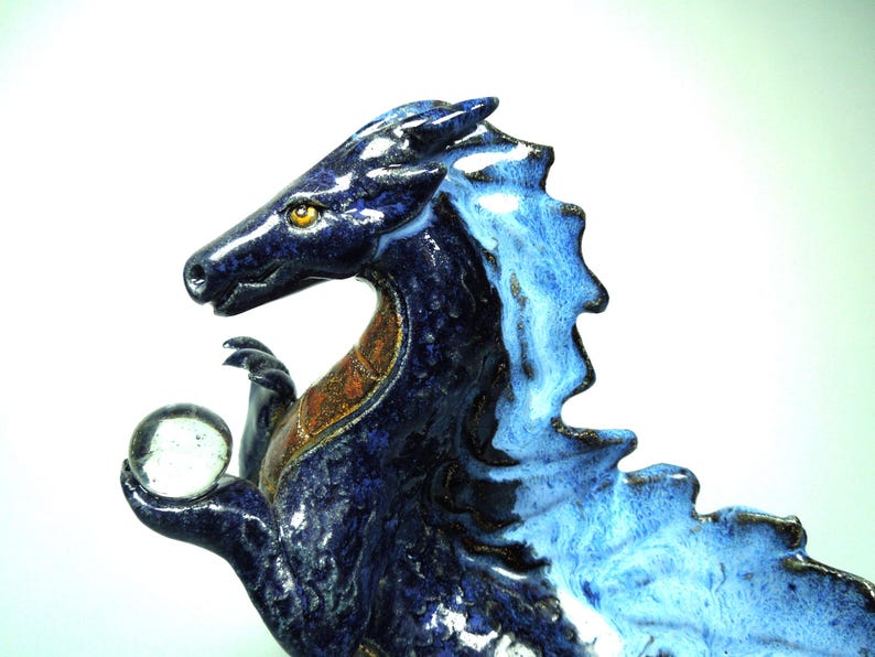 May include: A ceramic dragon figurine in shades of blue, holding a clear glass orb. The dragon has a detailed, textured surface with a contrasting orange-brown chest. The figurine is a decorative item.