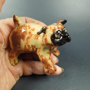 Puede incluir: Una peque&ntilde;a figura de cer&aacute;mica de un perro pug con un acabado brillante. El cuerpo es de color crema claro con manchas marrones moteadas, mientras que la cara y las orejas est&aacute;n pintadas con un esmalte negro s&oacute;lido y brillante.