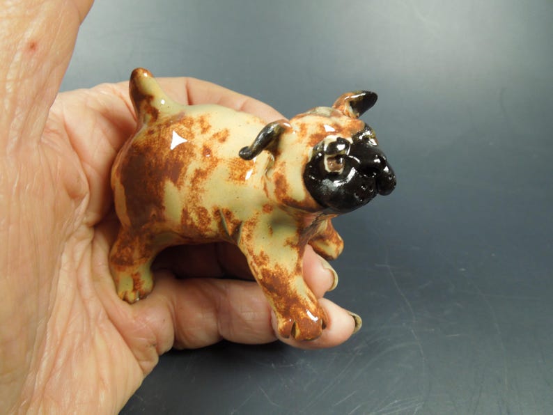 Puede incluir: Una peque&ntilde;a figura de cer&aacute;mica de un perro pug con un acabado brillante. El cuerpo es de color crema claro con manchas marrones moteadas, mientras que la cara y las orejas est&aacute;n pintadas con un esmalte negro s&oacute;lido y brillante.