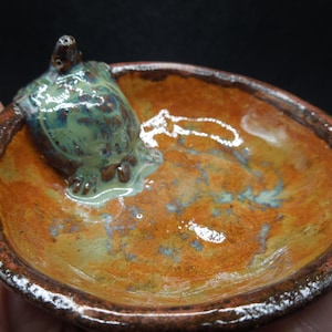 May include: A small ceramic dish with a glazed, orange-brown finish. The dish has a raised rim and a turtle figurine is sitting inside the dish. The turtle is glazed in shades of green and blue.