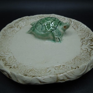 May include: A ceramic plate with a raised, textured border and a central, smooth area. A green turtle figurine with red eyes is centered on the plate. The border has a detailed, leaf-like pattern. The plate is off-white with a glossy finish.