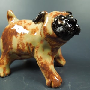 May include: A ceramic figurine of a pug dog with a glossy finish. The body is a light tan color with mottled brown patches throughout. The face, ears, and muzzle are painted in a solid, dark black glaze, giving the dog a distinct appearance.