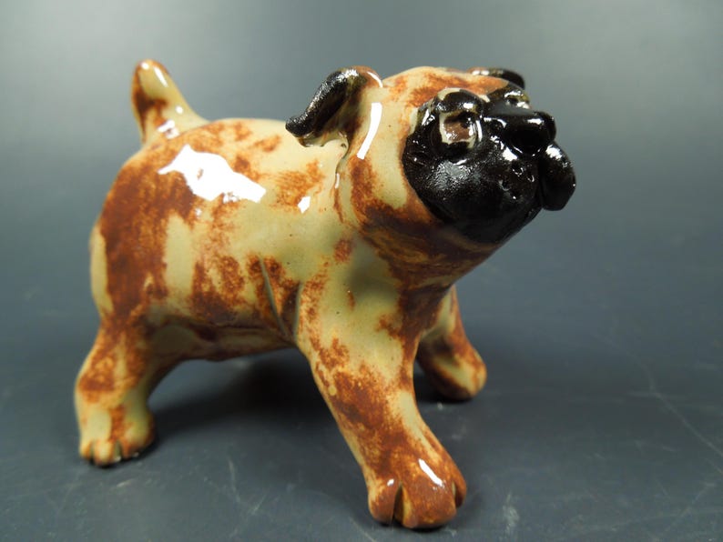 Puede incluir: Una figura de cer&aacute;mica de un perro pug con un acabado brillante. El cuerpo es de color beige claro con manchas marrones moteadas. La cara, las orejas y el hocico est&aacute;n pintados con un esmalte negro s&oacute;lido y oscuro, d&aacute;ndole un aspecto distintivo.
