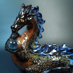May include: A ceramic dragon sculpture in shades of black, brown, and blue. The dragon holds a blue crystal ball. The sculpture has a textured surface and is posed in a dynamic, mythical stance.