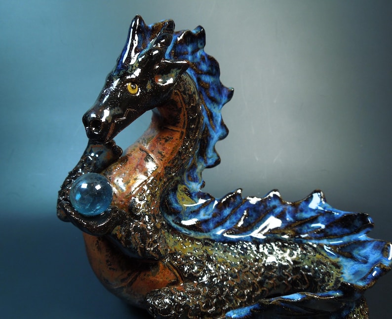 K&ouml;nnte beinhalten: Eine Keramikdrachenskulptur in Schwarz, Braun und Blau. Der Drache h&auml;lt eine blaue Kristallkugel. Die Skulptur hat eine strukturierte Oberfl&auml;che und ist in einer dynamischen, mythischen Haltung dargestellt.