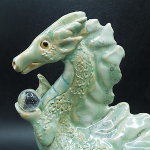 May include: A ceramic dragon sculpture in shades of green and tan, holding a clear crystal ball. The dragon has detailed scales and a textured surface. The sculpture has a whimsical, fantasy theme.