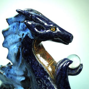 May include: A ceramic dragon figurine in shades of blue and brown, holding a clear glass orb. The dragon has a detailed, textured surface with yellow eyes. The figurine is a decorative item.